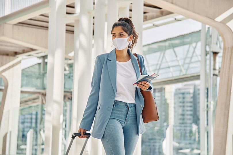 Mulher em aeroporto com máscara, bagagem e bilhetes de passagem.