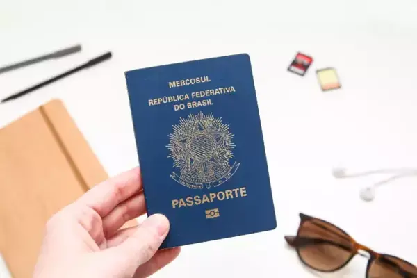 Pessoa segurando passaporte brasileiro ao lado de carteira e óculos de sol