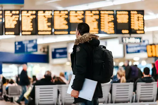 Homem em aeroporto segurando notebook enquanto olha o painel de voos.
