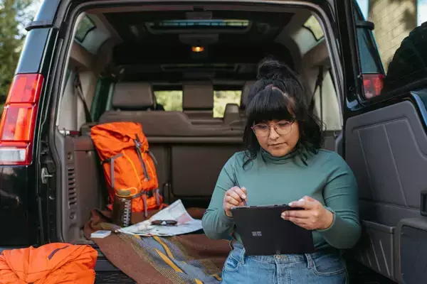 Foto de uma pessoa utilizando um tablet em frente ao carro, com a mala do carro aberta e algumas bagagens dentro.