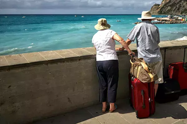 Foto de um casal de pessoas idosas admirando a paisagem de uma praia, ao lado de malas de viagem.