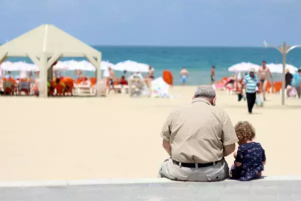 Foto de uma criança sentada na orla de uma praia com seu avô.