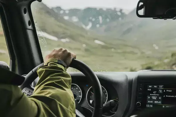 Foto de uma pessoa dentro do carro, dirigindo em direção à algumas montanhas, com um clima chuvoso.