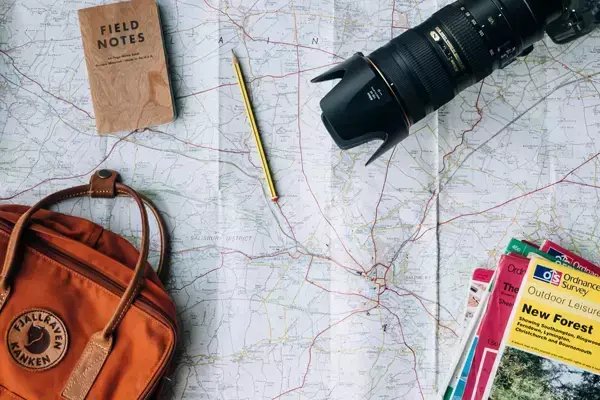 Imagem de um mapa com alguns itens típicos de viagem em cima dele: um bloco de notas, um lápis, uma câmera fotográfica, uma bolsa e livros sobre a natureza.