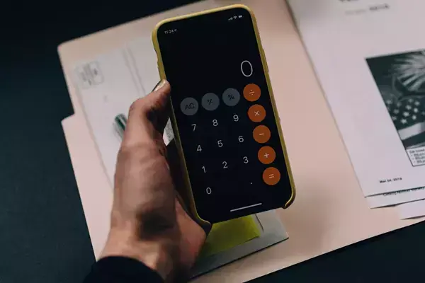 Foto da mão de uma pessoa segurando um celular com a calculadora aberta em cima de alguns documentos.