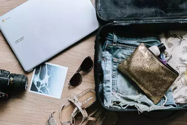 Foto de uma mala aberta, com roupas e uma bolsinha com produtos de limpeza. Do lado de fora da mala, vemos um par de sandálias, um notebook, um óculos de sol, uma câmera fotográfica e uma fotografia.