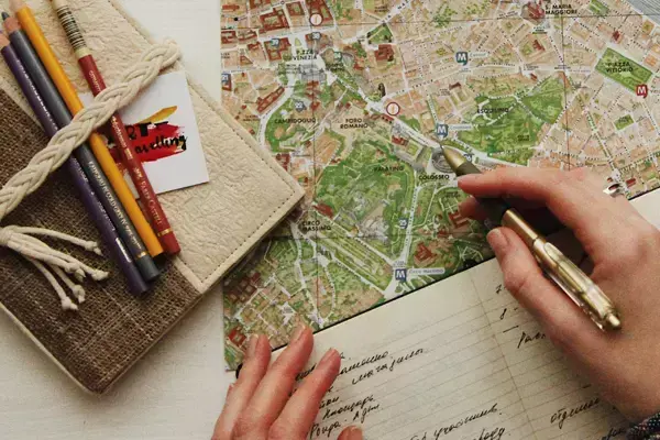 Foto de uma pessoa segurando uma caneta em cima de um caderno aberto e um mapa, com alguns objetos do lado.