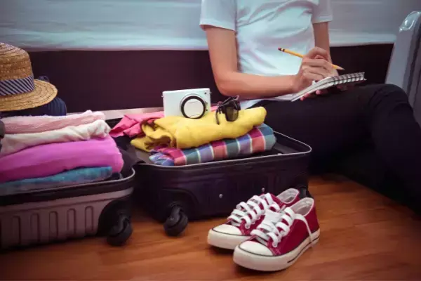 mulher sentada com caderneta e lápis na mão, ao lado de mala e itens de viagem