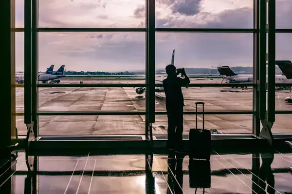 Homem em aeroporto com mala de bordo ao lado enquanto tira foto de avião com celular