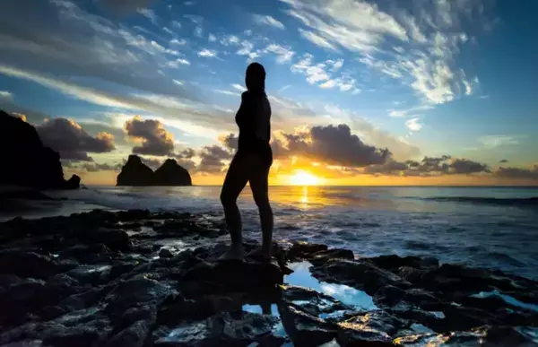 A sobra de uma mulher em cima das rochas perto do mar. Ao fundo, o sol se pondo.