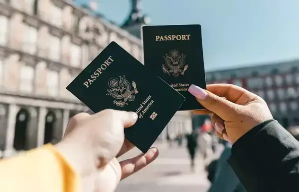 Duas pessoas tirando foto do passaporte delas, ao fundo um monumento histórico.