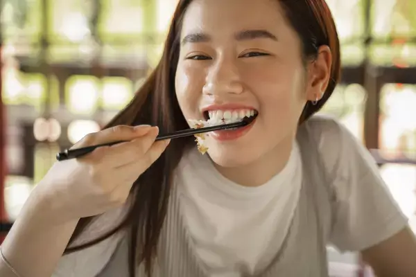 Foto frontal de uma mulher sorridente levando um macarrão chinês à boca.