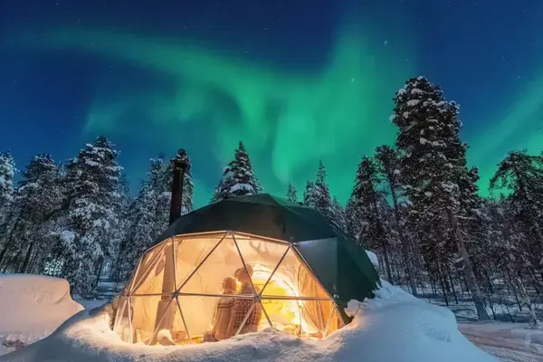 Casal dentro de cabana transparente vendo a Aurora Boreal no iluminando o céu de Lapônia