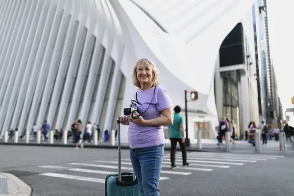 Senhora segurando câmera fotográfica e mala de viagem em frente a atração turística