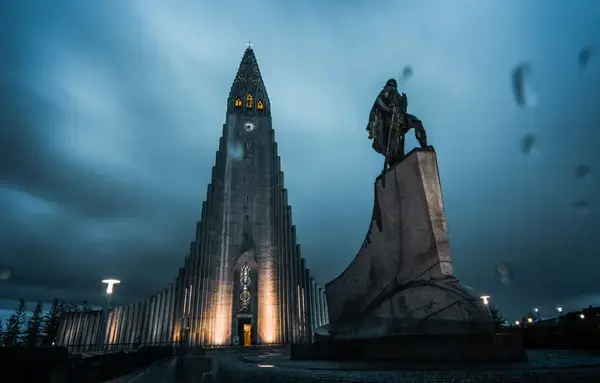 foto de um edifício alto e iluminado com o céu azul escuro e uma estatua em frente ao edificil