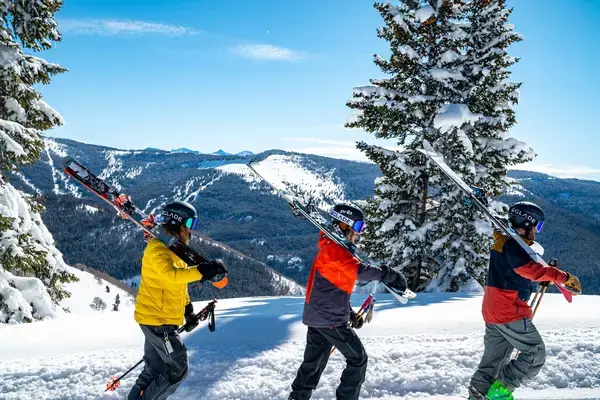 3 pessoas caminhando em fila na neve carregando equipamentos de esportes de inverno