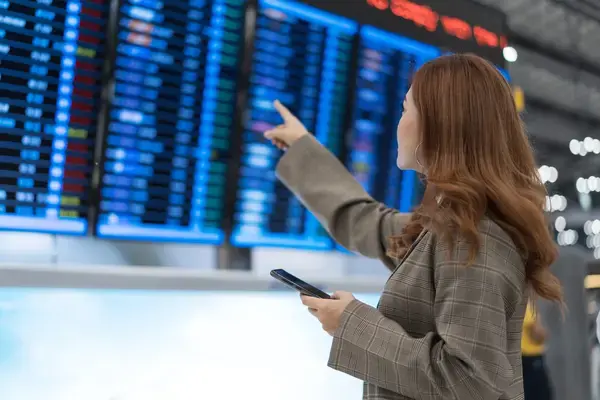 Mulher com celular na mão conferindo voo em painel de aeroporto