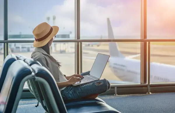 Mulher usando notebook enquanto observa aviões em aeroporto (Fonte: Adobe Stock)