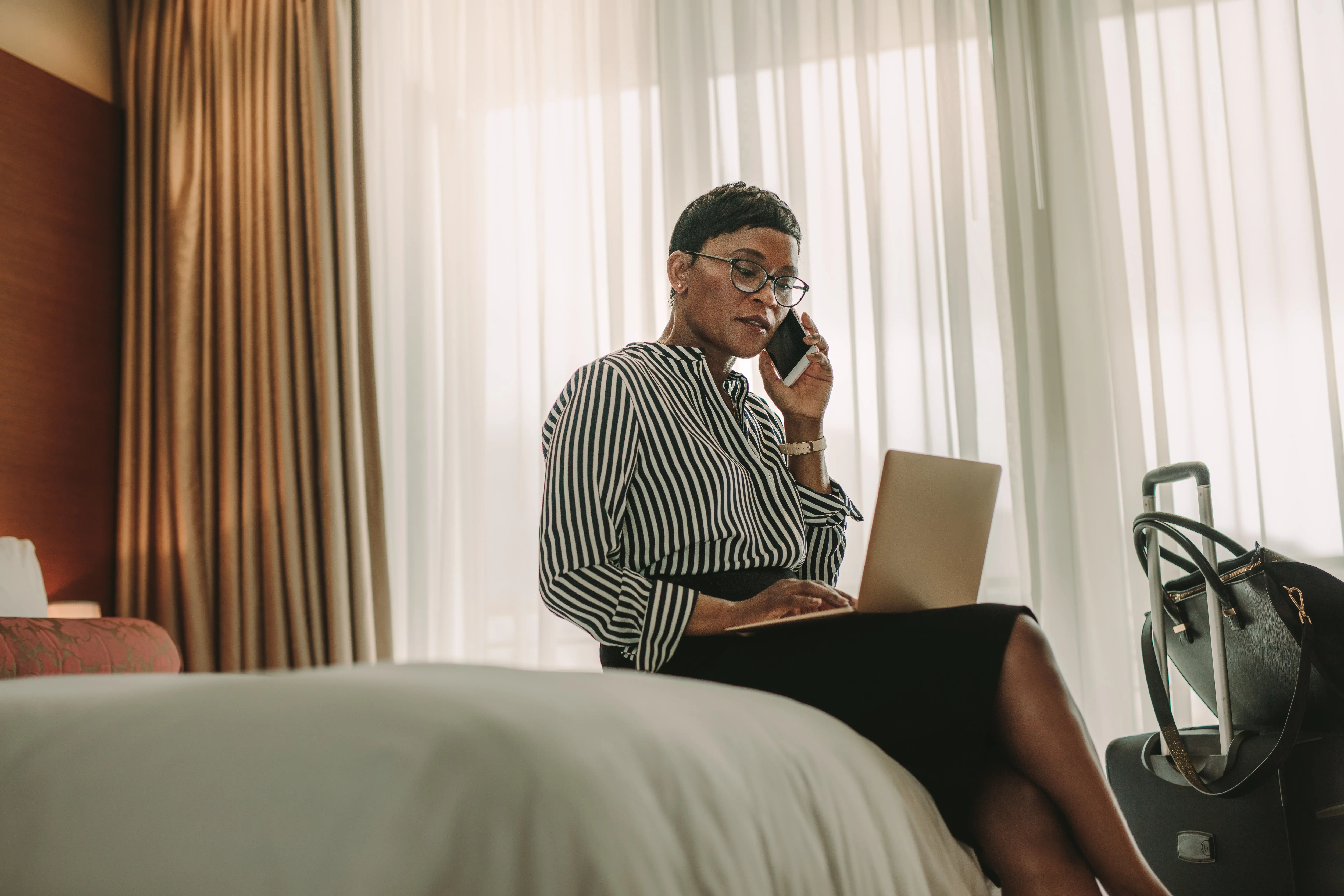 Mulher sentada na cama de hotel com notebook no colo e falando ao telefone (Fonte: Adobe Stock)