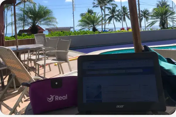 Computador e bolsinha da Real em primeiro plano, com o céu azul ao fundo.