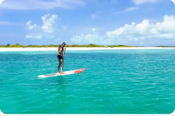 homem em pé em uma prancha sob as aguas azuis de los roques, com uma montanha ao fundo e areias brancas em primeiro plano