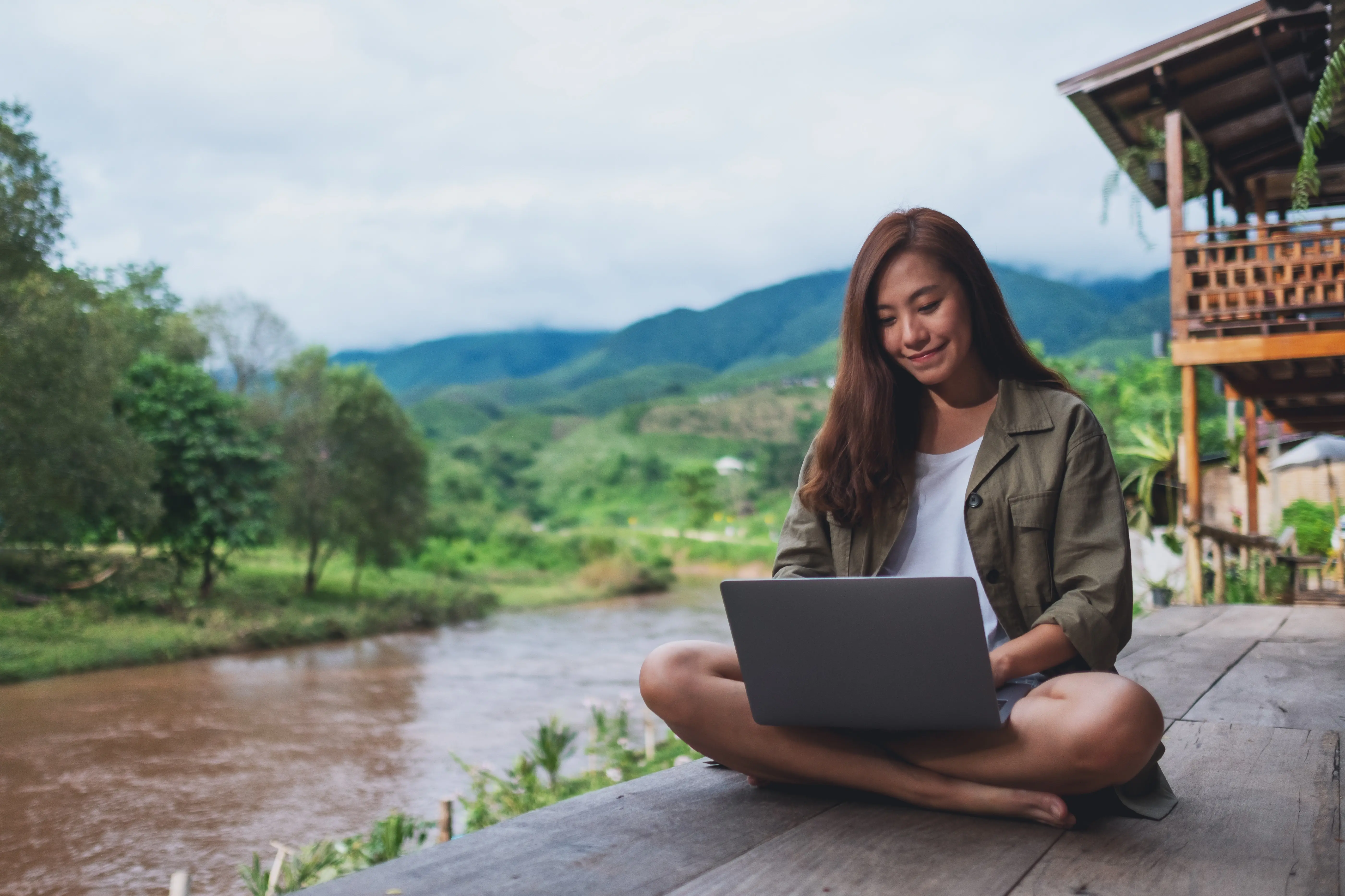 Mulher trabalhando no notebook em deck ao lado de rio entre montanhas (Fonte: Adobe Stock)
