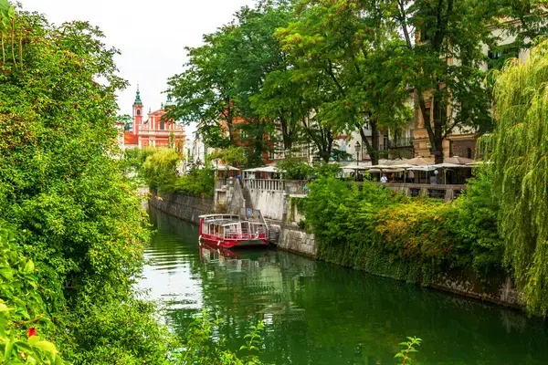 Barco turístico navegando pelo rio Ljubljanica entre árvores e cafés