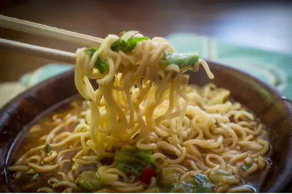 Close-up de hashis suspendendo uma porção de macarrão (noodles) de uma tigela de sopa com vegetais verdes.