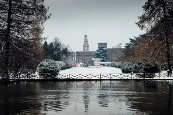 Parque em Milão coberto de neve com lago em primeiro plano e o Castello Sforzesco ao fundo durante o inverno italiano.