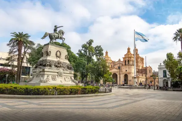 Praça central de Córdoba na Argentina com monumento a General San Martín e a catedral ao fundo, cercada por árvores, palmeiras e céu parcialmente nublado.