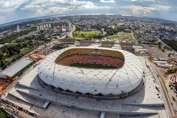 Vista aérea da Arena da Amazônia em Manaus, estádio com cobertura branca e formato circular, rodeado pela cidade e áreas verdes ao redor.