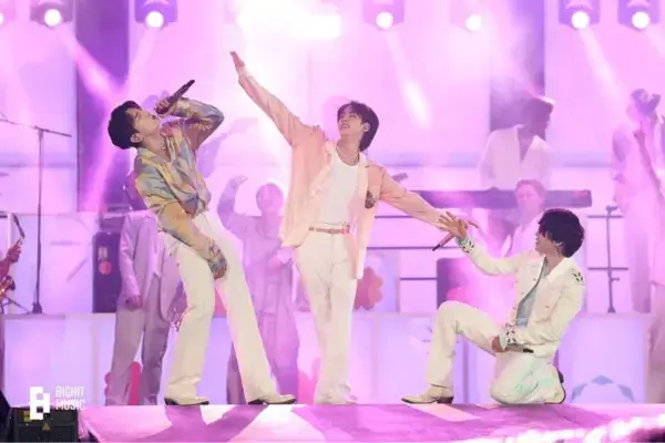 Jungkook, Jin e Jimin ao vivo em palco, com iluminação rosa e cenário vibrante, apresentando coreografia e cantando para fãs.