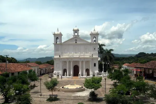 Fachada frontal da Igreja Santa Lucía em Suchitoto, uma construção colonial branca com duas torres sineiras e um relógio central. A igreja está situada no fundo de uma praça ampla com piso de pedra, árvores verdes espalhadas e cercada por casas de telhado de barro vermelho, sob um céu azul com nuvens e montanhas distantes.