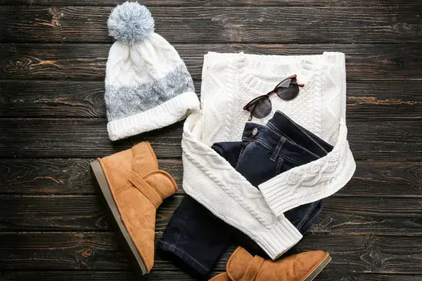 Conjunto de roupas de inverno com suéter, gorro, calça jeans, botas e óculos sobre superfície de madeira.