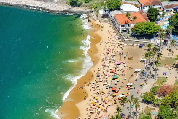 Vista aérea de praia movimentada com banhistas, guarda-sóis e mar verde.