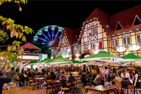 Foto noturna da Oktoberfest em Blumenau, Santa Catarina. Pessoas se reúnem em mesas ao ar livre sob guarda-sóis com a marca Spaten, em frente a prédios em estilo enxaimel. Ao fundo, uma roda-gigante iluminada completa o cenário da festa.
