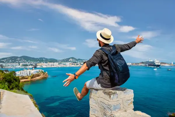 Homem sentado em um muro à beira-mar, de braços abertos, apreciando a vista de uma cidade costeira.