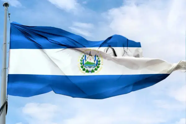 Bandeira de El Salvador tremulando em um mastro sob o céu azul com nuvens brancas. O estandarte possui três faixas horizontais, sendo duas azuis nas extremidades e uma branca no centro, onde fica o brasão de armas. No canto superior direito, a logo roxa da Real Seguro Viagem.