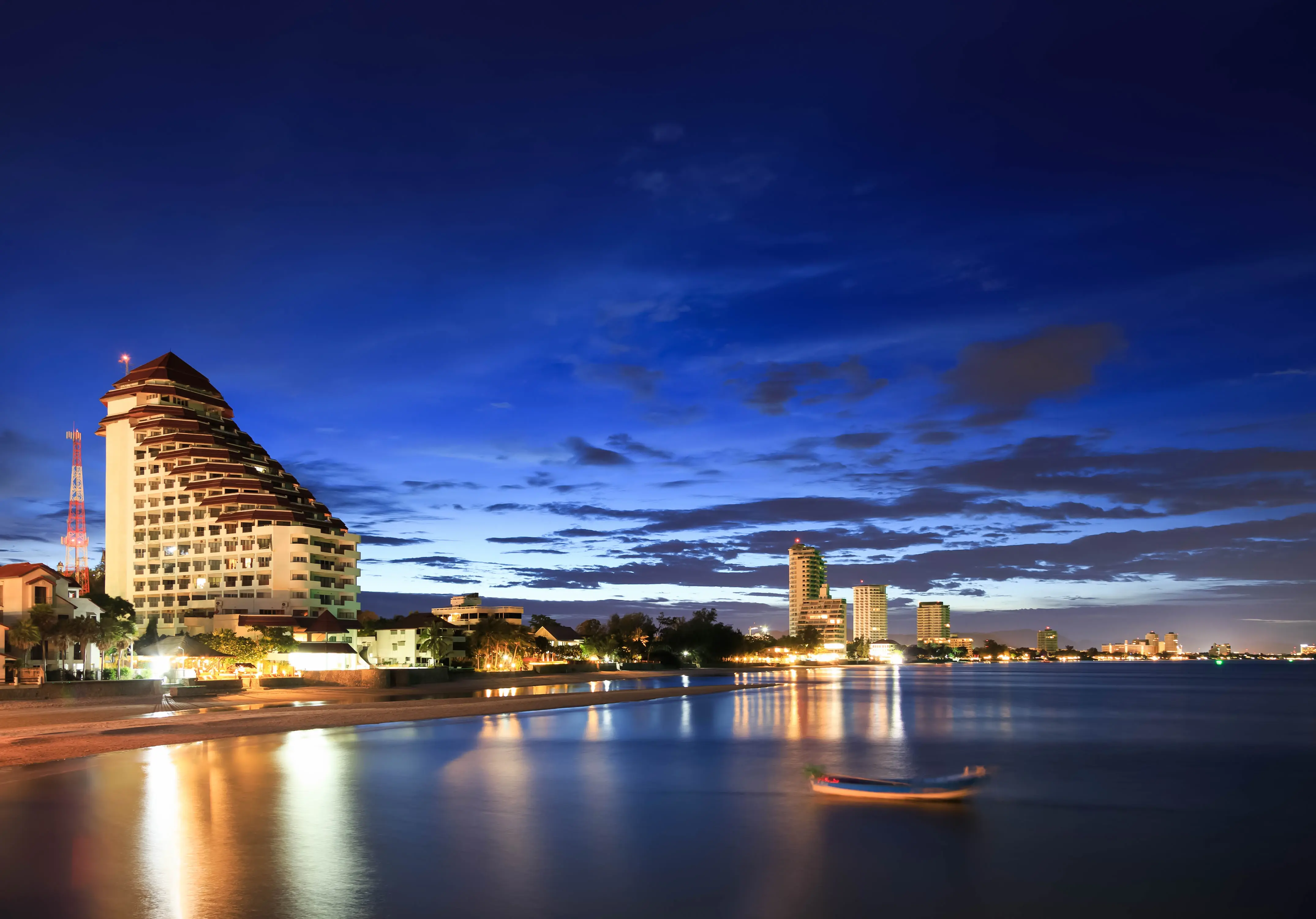 Vista panorâmica da praia de Hua Hin na Tailândia durante o crepúsculo. À esquerda, destaca-se um grande hotel iluminado com arquitetura em degraus. O mar calmo reflete as luzes da cidade e do céu azul profundo, com um pequeno barco solitário em primeiro plano.