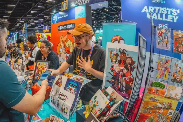 Um estande de artistas na CCXP, com um ilustrador interagindo com um visitante enquanto exibe suas obras em uma mesa. Ao fundo, outros artistas estão em seus estandes, e várias ilustrações coloridas estão expostas.