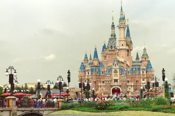Vista frontal do majestoso castelo da Disney em Xangai, com suas torres azuis e douradas sob um céu claro. Em primeiro plano, jardins floridos, postes de luz clássicos e visitantes caminhando pelas pontas e áreas comuns do parque.
