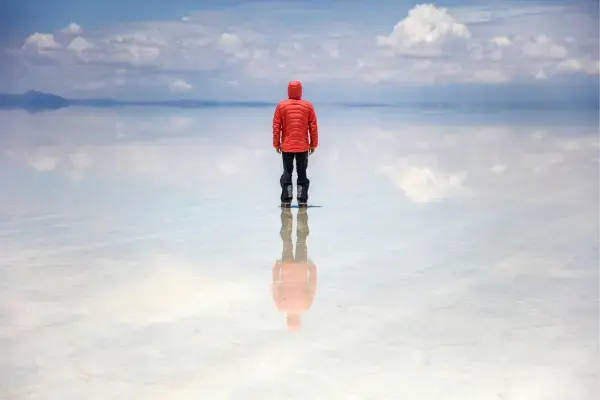 Homem de jaqueta vermelha refletido no Salar de Uyuni na Bolívia com céu de nuvens