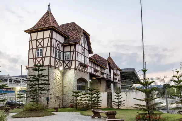 Construção em estilo enxaimel com torres e telhado inclinado, típica da arquitetura germânica, no Vale Europeu de Santa Catarina. O prédio é cercado por jardim com bancos e pinheiros, com céu claro ao entardecer.