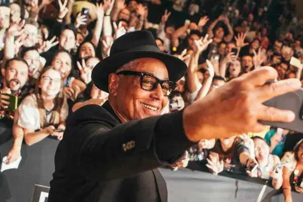 Ator Giancarlo Esposito tirando uma selfie com fãs ao fundo, todos sorrindo e acenando