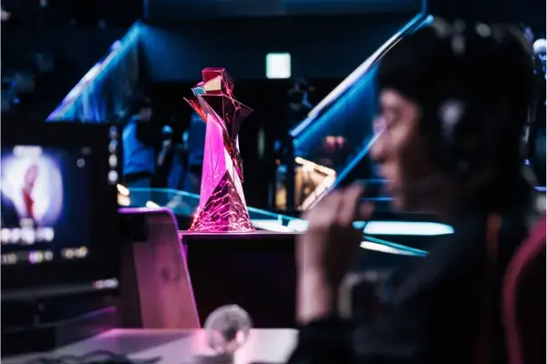 Troféu internacional de League of Legends iluminado no palco durante partida de esports, com jogadores e computadores ao fundo em uma arena competitiva.