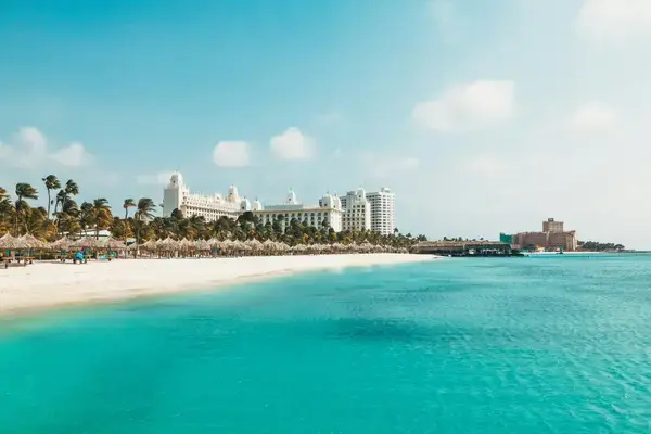 Mar azul cristalino e faixa de areia clara com palmeiras e hotéis ao fundo em Aruba.