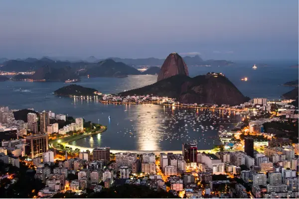 Vista aérea do Rio de Janeiro ao entardecer, com o Pão de Açúcar ao centro, a Baía de Guanabara refletindo as luzes da cidade e a arquitetura urbana ao redor.