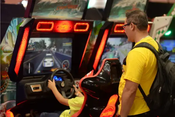 Uma criança jogando em um simulador de corrida enquanto um adulto a observa ao lado. Ambos usam camiseta amarela, e o ambiente tem iluminação de máquinas de fliperama.