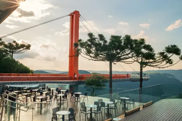 Deck com mesas do Skyglass Canela com vista para o vale e araucárias, passarela suspensa ao fundo