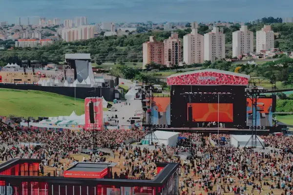Área do Lollapalooza Brasil durante o dia, com palcos, estandes, público circulando e prédios residenciais ao fundo em São Paulo.